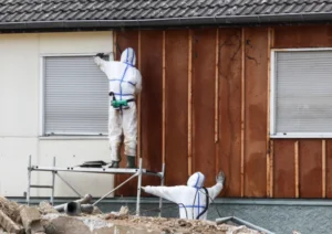 Specialized remediation workers in full hazard suits stripping exterior siding from a building during an abatement or restoration project.